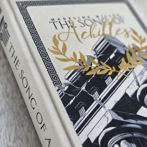 May include: A hardcover book titled "The Song of Achilles" by Madeline Miller. The book has a cream-colored cover with gold lettering and a black and white illustration of classical architecture. A gold laurel wreath accents the cover.