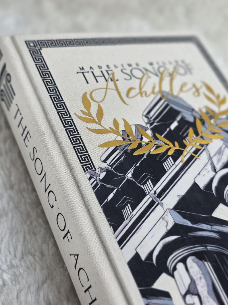 May include: A hardcover book titled "The Song of Achilles" by Madeline Miller. The book has a cream-colored cover with gold lettering and a black and white illustration of classical architecture. A gold laurel wreath accents the cover.