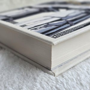 May include: A hardcover book with a white linen cover. The book's cover features a geometric design in shades of gray and black. The pages are a light cream color, and the book is resting on a white, fluffy surface.