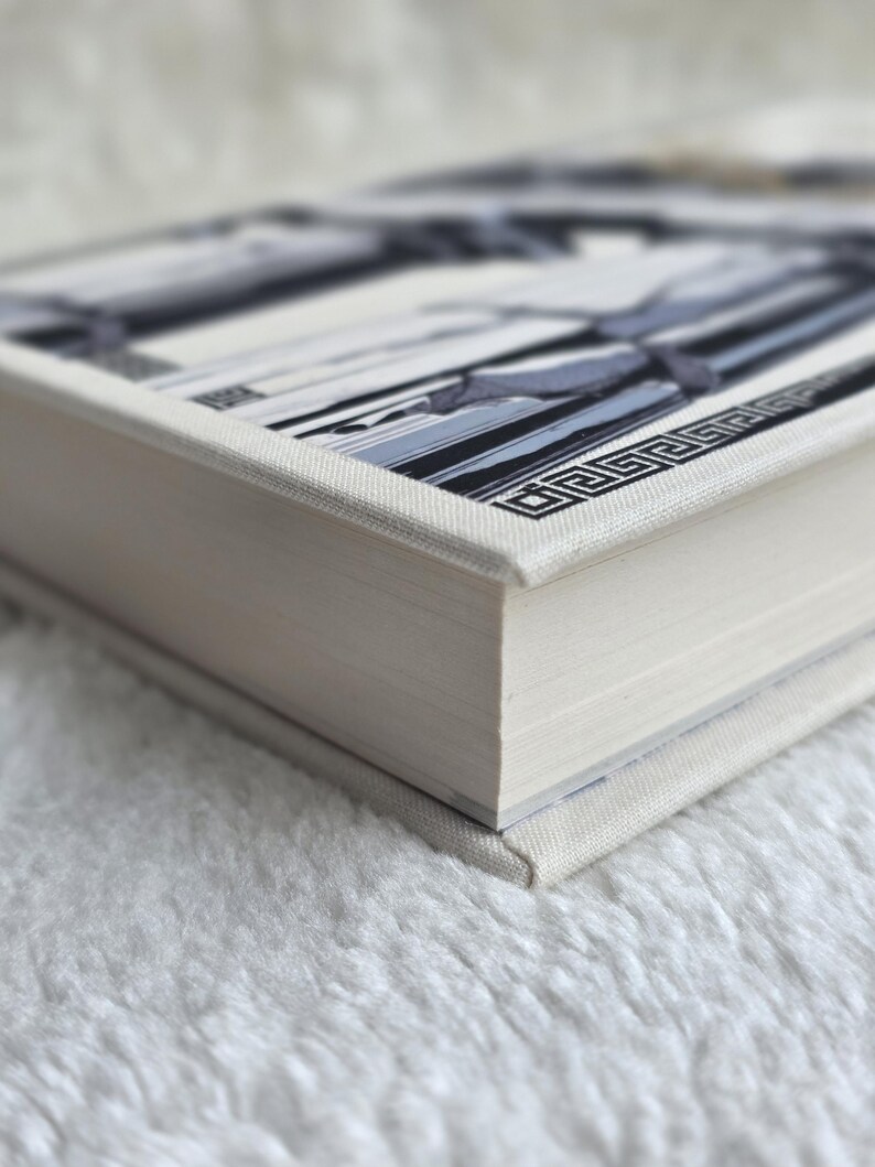 May include: A hardcover book with a white linen cover. The book's cover features a geometric design in shades of gray and black. The pages are a light cream color, and the book is resting on a white, fluffy surface.