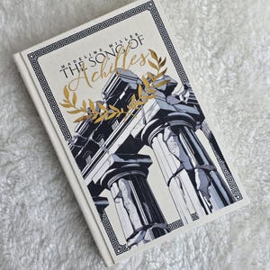May include: A hardcover book titled "The Song of Achilles" by Madeline Miller. The cover features a black and white illustration of classical columns with gold accents and text. The book is on a white, textured surface.