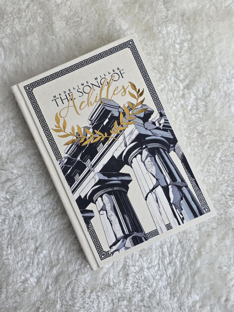 May include: A hardcover book titled "The Song of Achilles" by Madeline Miller. The cover features a black and white illustration of classical columns with gold accents and text. The book is on a white, textured surface.