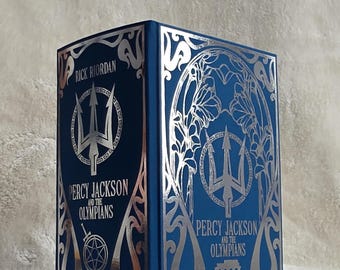 May include: A hardcover book with a blue cover and silver foil accents. The book is titled "Percy Jackson and the Olympians" by Rick Riordan. The cover features a trident and decorative floral designs.