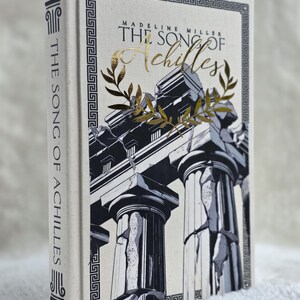 May include: Hardcover book titled "The Song of Achilles" by Madeline Miller. The book cover features a black and white illustration of Greek columns with gold accents and text. The spine displays the title vertically.