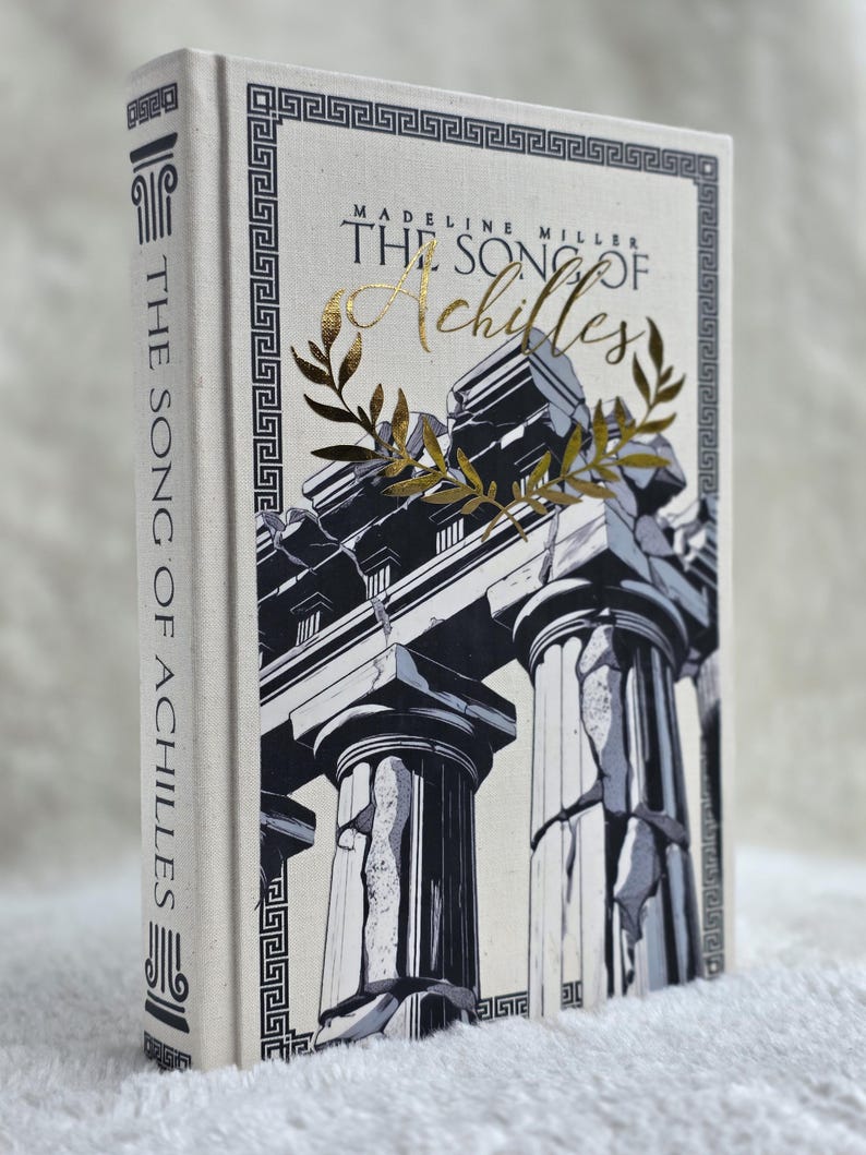 May include: Hardcover book titled "The Song of Achilles" by Madeline Miller. The book cover features a black and white illustration of Greek columns with gold accents and text. The spine displays the title vertically.