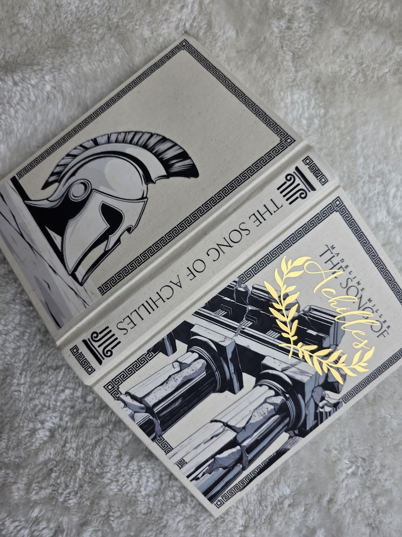 May include: Open book titled "The Song of Achilles" with a cream-colored cover. The cover features a black and white illustration of a Greek helmet and architectural columns. Gold lettering and a laurel wreath accent the title.