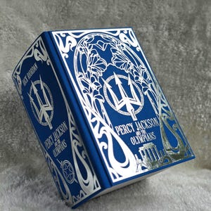 May include: A blue hardback book titled Percy Jackson and the Olympians by Rick Riordan. The cover features intricate silver foil detailing, including a central trident emblem, floral patterns, and a silhouette of a Greek temple at the bottom.