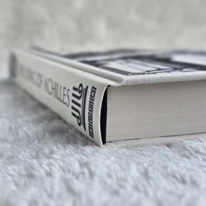 May include: A hardcover book titled "The King of Achilles" with a white cover and black text. The book's spine features stylized text and a decorative design. The book's pages are white, and the edges are black.