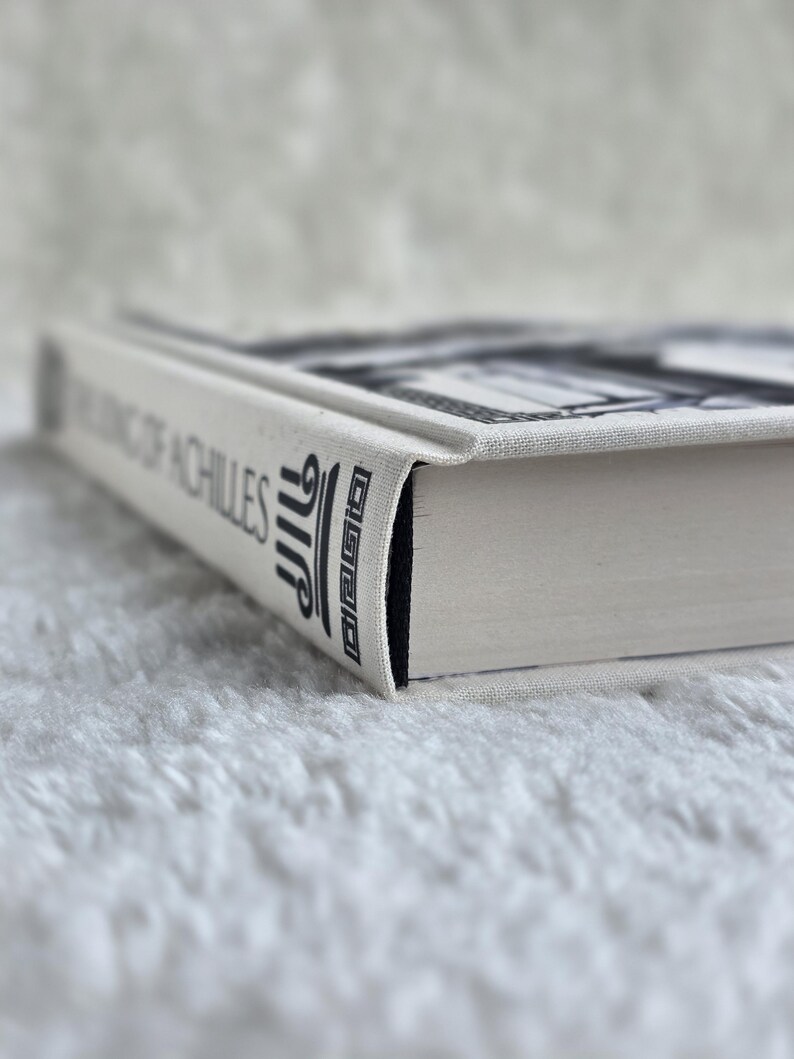 May include: A hardcover book titled "The King of Achilles" with a white cover and black text. The book's spine features stylized text and a decorative design. The book's pages are white, and the edges are black.