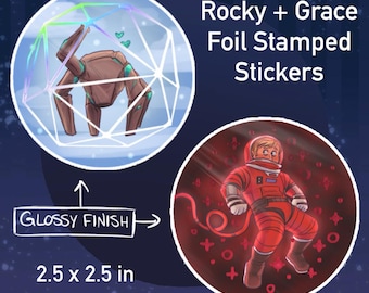 Op de afbeelding: Twee ronde stickers met een glanzende afwerking. Eén toont een bruine robot in een geometrische vorm, de andere een astronaut in een rood pak. De tekst luidt "Rocky + Grace Foil Stamped Stickers". Elke sticker is 6,35 x 6,35 cm groot.