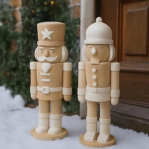 Puede incluir: Dos cascanueces de madera, uno con una estrella en el sombrero y el otro con un sombrero redondo. Ambos están pintados en colores marrón claro y crema, de pie sobre una superficie nevada. Son decoraciones navideñas clásicas.