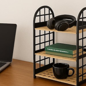 May include: A black metal three-tier shelf unit with a grid design. The shelves hold a pair of black headphones, two green books, and a black mug. A laptop sits to the left on a wooden desk.