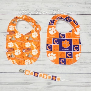 May include: Two orange baby bibs and a pacifier clip with a Clemson Tigers theme. One bib features paw prints and football helmets, the other has a checkerboard pattern with the Clemson logo. The pacifier clip has a matching design.