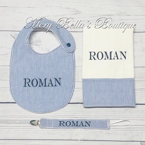 May include: A baby gift set featuring a bib, burp cloth, and pacifier clip. The items are white and blue striped with the name "ROMAN" embroidered in navy blue. The bib has a snap closure, and the pacifier clip has a white ribbon.