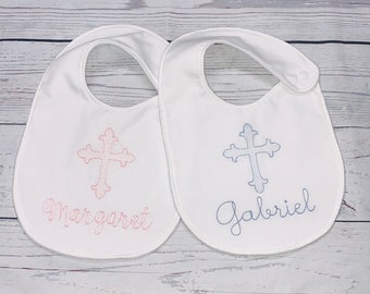May include: Two white bibs with embroidered crosses and names. The bib on the left has a pink cross and the name "Margaret" embroidered in pink. The bib on the right has a blue cross and the name "Gabriel" embroidered in blue.