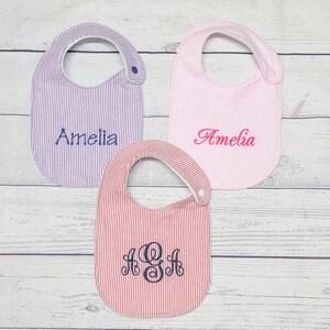 May include: Three baby bibs with striped patterns. One bib is purple and white, one is pink and white, and one is red and white. The bibs are embroidered with the name "Amelia" and the monogram "A&H".