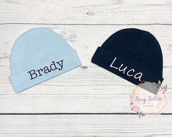 May include: Two baby hats, one light blue with the name "Brady" embroidered on it and one dark blue with the name "Luca" embroidered on it.