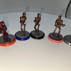 Puede incluir: Figuras en miniatura de droides de combate de color bronce sobre bases grises con bordes de colores. Las bases tienen bordes naranja, azul, negro y rosa. Algunos droides sostienen armas. Estas figuras son probablemente para juegos de mesa o exhibición.