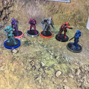 Puede incluir: Una colección de figuras en miniatura, cada una representando un personaje en varios colores, incluyendo turquesa, morado, rosa, rojo y azul. Las figuras están sobre bases circulares de diferentes colores, sobre una superficie texturizada marrón y verde.