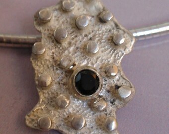 May include: A silver pendant with a textured surface and a black gemstone in the center. The pendant is attached to a silver chain.