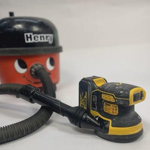 May include: A red and black Henry vacuum cleaner with a smiling face and the name "Henry" on top. A black and yellow DeWalt sander is connected to the vacuum cleaner with a black hose.