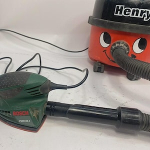 May include: A red and black Henry vacuum cleaner with a smiling face design is connected to a green and black Bosch PSM 100 A sander. The sander is connected to the vacuum cleaner with a black hose. The vacuum cleaner has the word "Henry" on it.