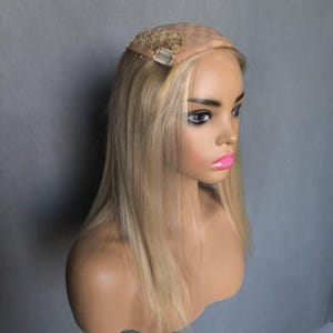 Op de afbeelding: Blonde pruik met steil, halflang haar, tentoongesteld op een mannequinhoofd. De pruik heeft een kanten voorkant en een kleine kam voor een veilige bevestiging. De haarkleur is een mix van lichte en medium blonde tinten.