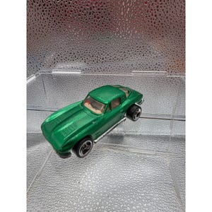 May include: A metallic green toy sports car featuring a sleek coupe body style, a tan interior, and black wheels with silver rims. The vehicle has a long bonnet with visible vents and a low-profile design, captured from a high-angle perspective.