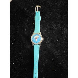 Puede incluir: Un reloj de pulsera con correa azul claro y caja circular de tono plateado. La esfera es azul claro con números negros y presenta una ilustración central del personaje Snoopy. Tiene manecillas rojas y un segundero de color plateado.