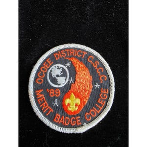 Può includere: Una toppa ricamata circolare con sfondo grigio scuro e bordo bianco. Presenta il testo arancione OCOEE DISTRICT C.S.C.C. MERIT BADGE COLLEGE, un globo, un giglio giallo, un disegno di cometa fiammeggiante e l'anno '89.
