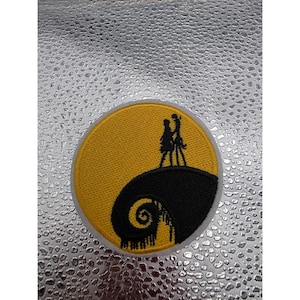 May include: A circular embroidered patch featuring a yellow moon background with a black silhouette of two figures standing on a curved, spiraling hill. The design depicts a scene with a dark, textured foreground and a bright, circular yellow backdrop.