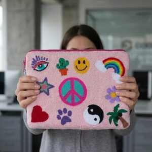 May include: A pink laptop sleeve with a variety of embroidered patches, including a rainbow, smiley face, peace sign, and heart. The sleeve has a zipper closure and a pink border. The design is colorful and whimsical.