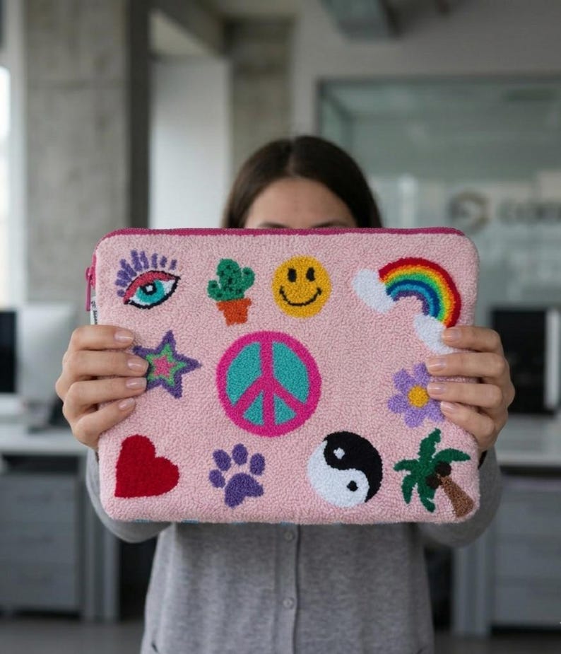May include: A pink laptop sleeve with a variety of embroidered patches, including a rainbow, smiley face, peace sign, and heart. The sleeve has a zipper closure and a pink border. The design is colorful and whimsical.