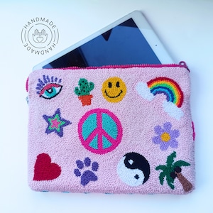 以下が含まれることがあります： ピンク色の手作りのタブレットケース。ファスナー開閉式。ハート、星、平和のシンボル、スマイリーフェイス、虹、目、足跡、花、サボテン、陰陽、ヤシの木の刺繍パッチで装飾されています。iPadが一部見えています。