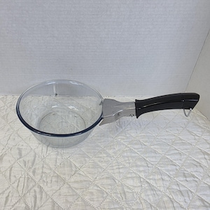 May include: A clear glass saucepan with a round, transparent bowl and a smooth, black plastic handle. The handle is attached to the glass body with a silver-coloured metal bracket and features a small metal loop at the end for hanging storage.