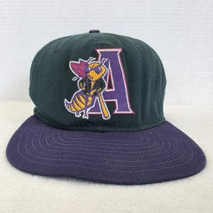 Può includere: Un berretto da baseball verde scuro con visiera viola. Il berretto presenta una lettera "A" viola e un'ape dei cartoni animati con una mazza da baseball. L'ape è gialla e nera con la testa viola. Il berretto è in stile baseball classico.