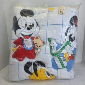 Puede incluir: Una almohada blanca para bebé con los personajes de dibujos animados Mickey Mouse y Pluto. Mickey lleva trajes rojos y azules, y Pluto es amarillo. La almohada está en una bolsa de plástico y tiene un patrón de cuadrícula.