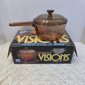 May include: A transparent amber-coloured glass saucepan with a matching lid, featuring a long handle. The saucepan sits on its original cardboard box, which displays the text VISIONS, 1-QT/1 L COVERED SAUCEPAN V-1, and COOKWARE THAT LETS YOU SEE WHAT'S COOKING.