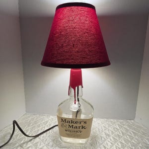 Könnte beinhalten: Eine einzigartige Lampe aus einer Maker's Mark Whiskey-Flasche. Sie hat einen roten Lampenschirm und ein rotes, wachsartiges Detail um den Flaschenhals. Das Etikett der Flasche ist sichtbar. Ein schwarzes Netzkabel erstreckt sich von der Basis.