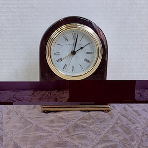 May include: A Howard Miller wooden clock with a gold-tone bezel and Roman numeral hour markers. The clock has a dark brown, arched wooden case and a white clock face. The clock is sitting on a gold-tone base.