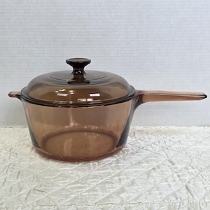 May include: A brown glass saucepan with a lid and a long handle. The saucepan is made of transparent glass, allowing visibility of the contents. The lid has a small, rounded knob. The handle extends to the right.