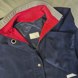 May include: Navy blue jacket with a red interior lining and a gray collar. The jacket has a front zipper, two pockets, and a button closure. The label inside the jacket reads "CBO".