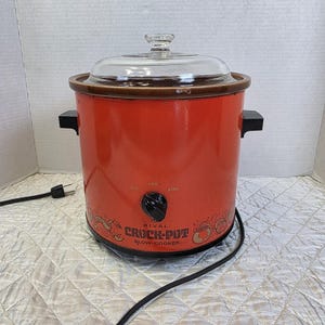 May include: A bright orange Rival Crock-Pot slow cooker with a clear glass lid and black side handles. The front features a black control knob with off, low, and high settings, along with decorative line art illustrations of kitchen pots.