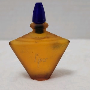 Puede incluir: Un frasco de perfume vintage de color ámbar con un tapón azul. El frasco tiene un diseño geométrico y las palabras "le jour" están grabadas en la parte delantera. El frasco tiene una forma única y un aspecto esmerilado.