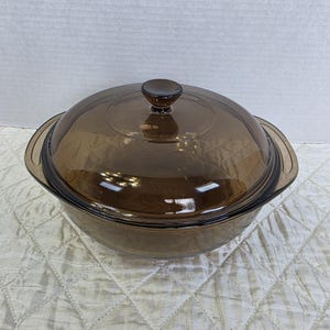 May include: A brown glass casserole dish with a matching lid. The dish has two handles and a round knob on top of the lid. The glass is transparent, allowing the contents to be seen. The dish is designed for cooking and serving.
