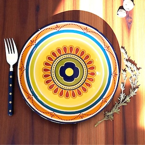 May include: A colorful ceramic plate with a floral design in the center. The plate features a yellow background with a blue, green, orange, and red pattern. A silver fork with a dark blue handle is to the left.