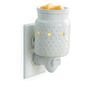 May include: A white ceramic wax warmer with a textured, dotted surface, plugged into a wall outlet. The warmer has a jar-like shape with a wide opening, filled with orange wax melts. Three small holes emit a warm, yellow glow.