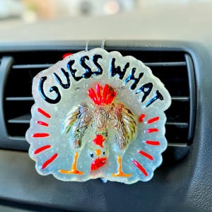 May include: A translucent, scalloped-edge air freshener featuring a glittery finish and the black text GUESS WHAT. A central graphic depicts a chicken upside down with red and orange accents, surrounded by small red dashes radiating outward.