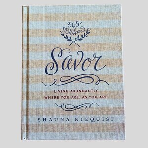 May include: A hardcover book titled "Savor" with a striped beige and white cover. The title is in elegant navy blue script, with the words "365 Devotions" above. The author's name, Shauna Niequist, is at the bottom.