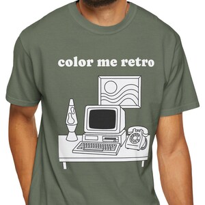 Puede incluir: Camiseta verde oliva con el texto "color me retro" sobre un gráfico blanco de una configuración de escritorio retro. El gráfico incluye una computadora, una lámpara de lava, un teléfono y una imagen enmarcada de un sol y olas.